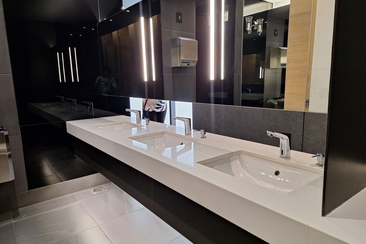 Modern commercial bathroom featuring a sleek triple-basin vanity with integrated white sinks, chrome sensor faucets, and large wall-to-wall mirrors with integrated lighting.