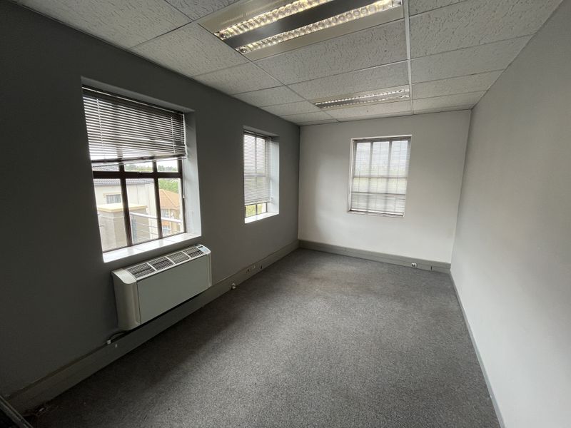 A vacant office space featuring grey carpeting, suspended ceiling with recessed lighting, and a wall-mounted HVAC unit with natural light from multiple windows.