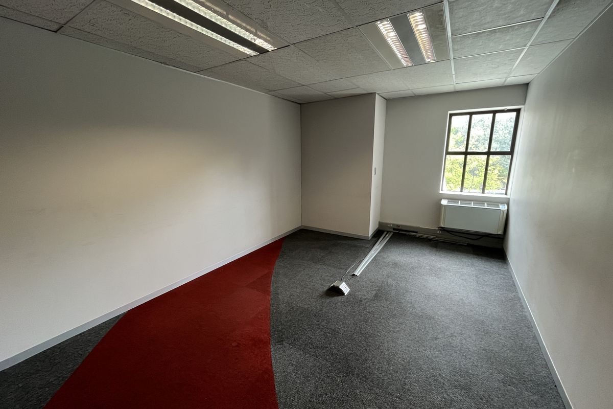 An empty office space featuring carpet tiles, a suspended ceiling with recessed lighting, and a window with an integrated HVAC unit.
