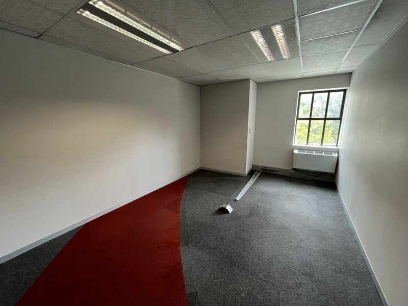 An empty office space featuring carpet tiles, a suspended ceiling with recessed lighting, and a window with an integrated HVAC unit.