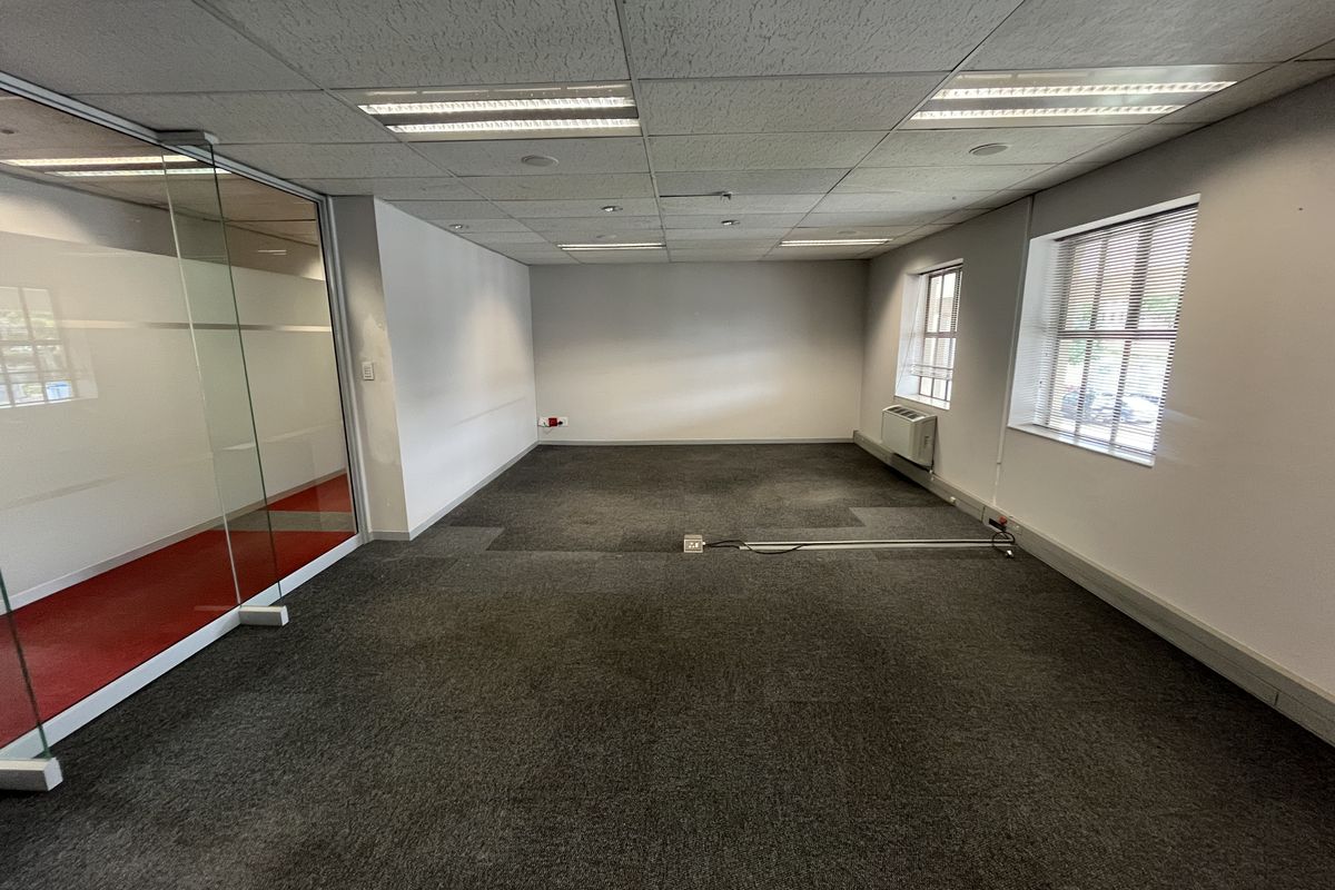 A vacant office space featuring carpeted flooring, a glass partition wall, suspended ceiling with recessed lighting, and wall-mounted climate control.