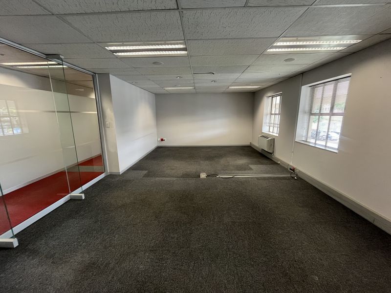 A vacant office space featuring carpeted flooring, a glass partition wall, suspended ceiling with recessed lighting, and wall-mounted climate control.