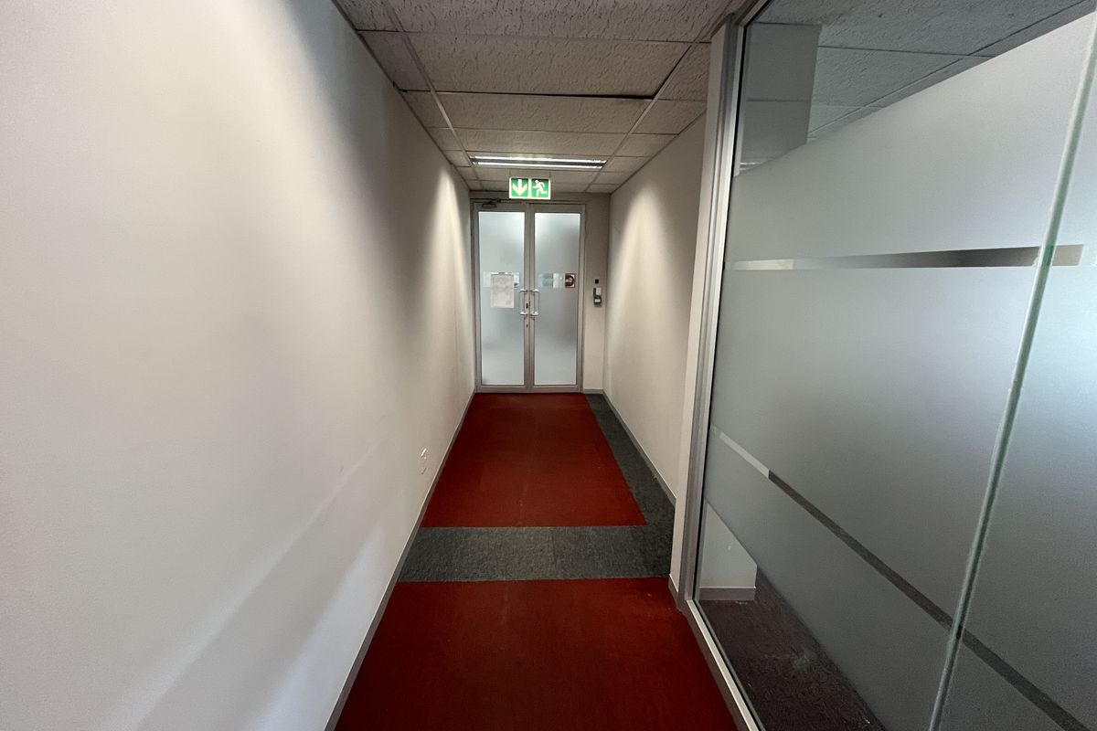 A functional office hallway featuring a suspended ceiling, frosted glass partitioning, and double glass doors leading to an emergency exit.