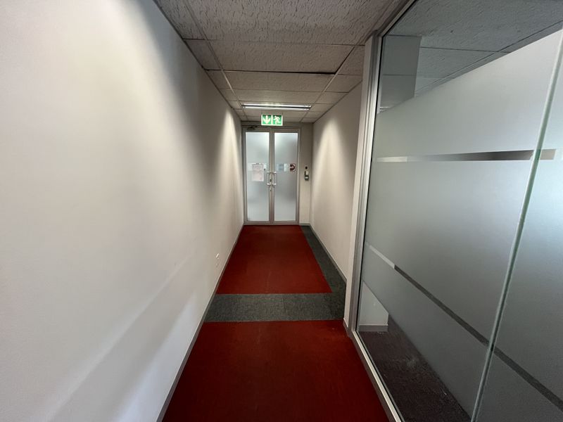 A functional office hallway featuring a suspended ceiling, frosted glass partitioning, and double glass doors leading to an emergency exit.