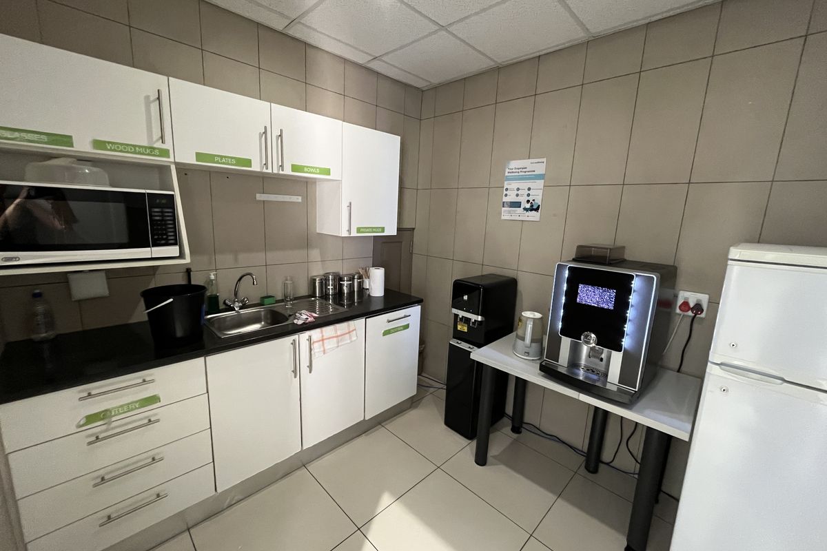 A functional office kitchenette featuring white cabinetry, dark countertops, a stainless steel sink, and tiled walls and flooring.