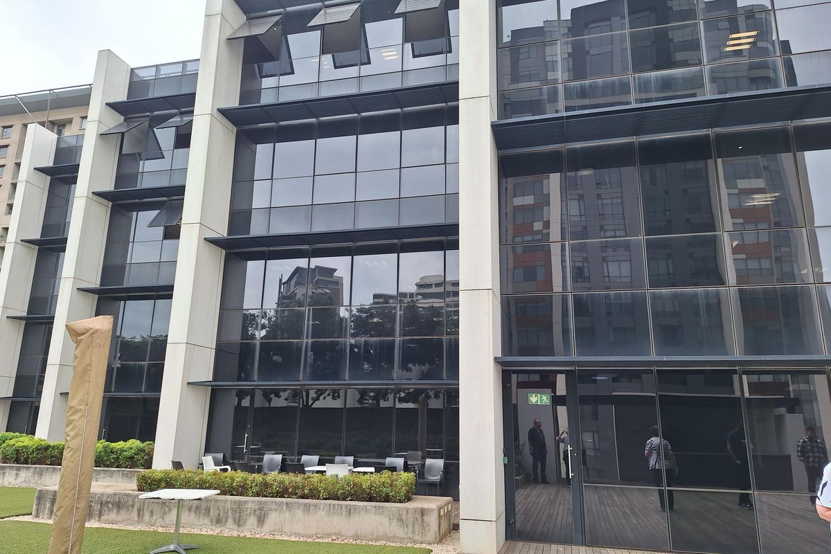 Modern office building exterior featuring a glass curtain wall facade, concrete structural columns, and an adjacent outdoor patio area with decking.