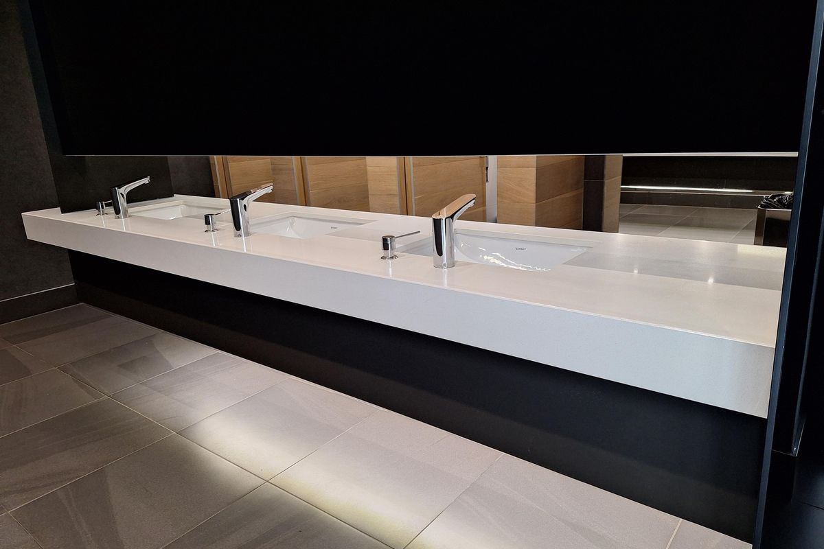 Modern office restroom featuring a sleek, wall-mounted white vanity with multiple integrated sinks, chrome faucets, and large mirror.