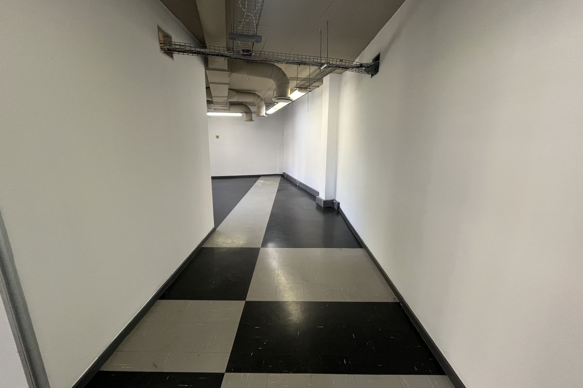 A vacant office hallway featuring exposed ceiling ducting, cable trays, and a black and grey checkered vinyl floor.