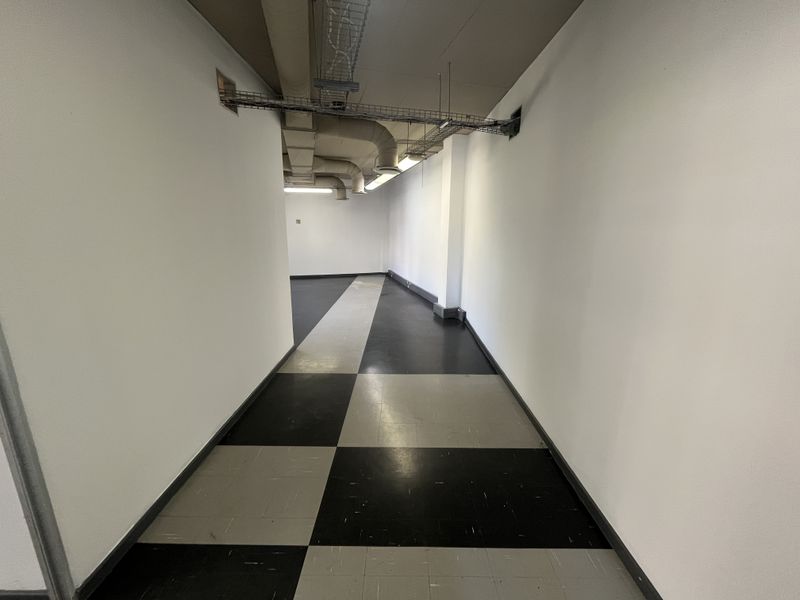 A vacant office hallway featuring exposed ceiling ducting, cable trays, and a black and grey checkered vinyl floor.