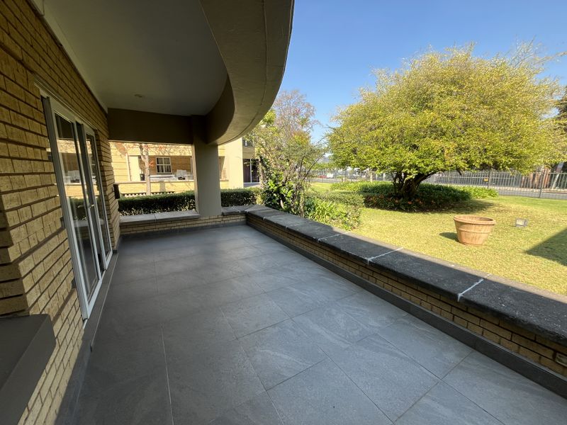 A spacious covered patio featuring tiled flooring, a brick facade, and a concrete ledge, overlooking a well-maintained garden area.