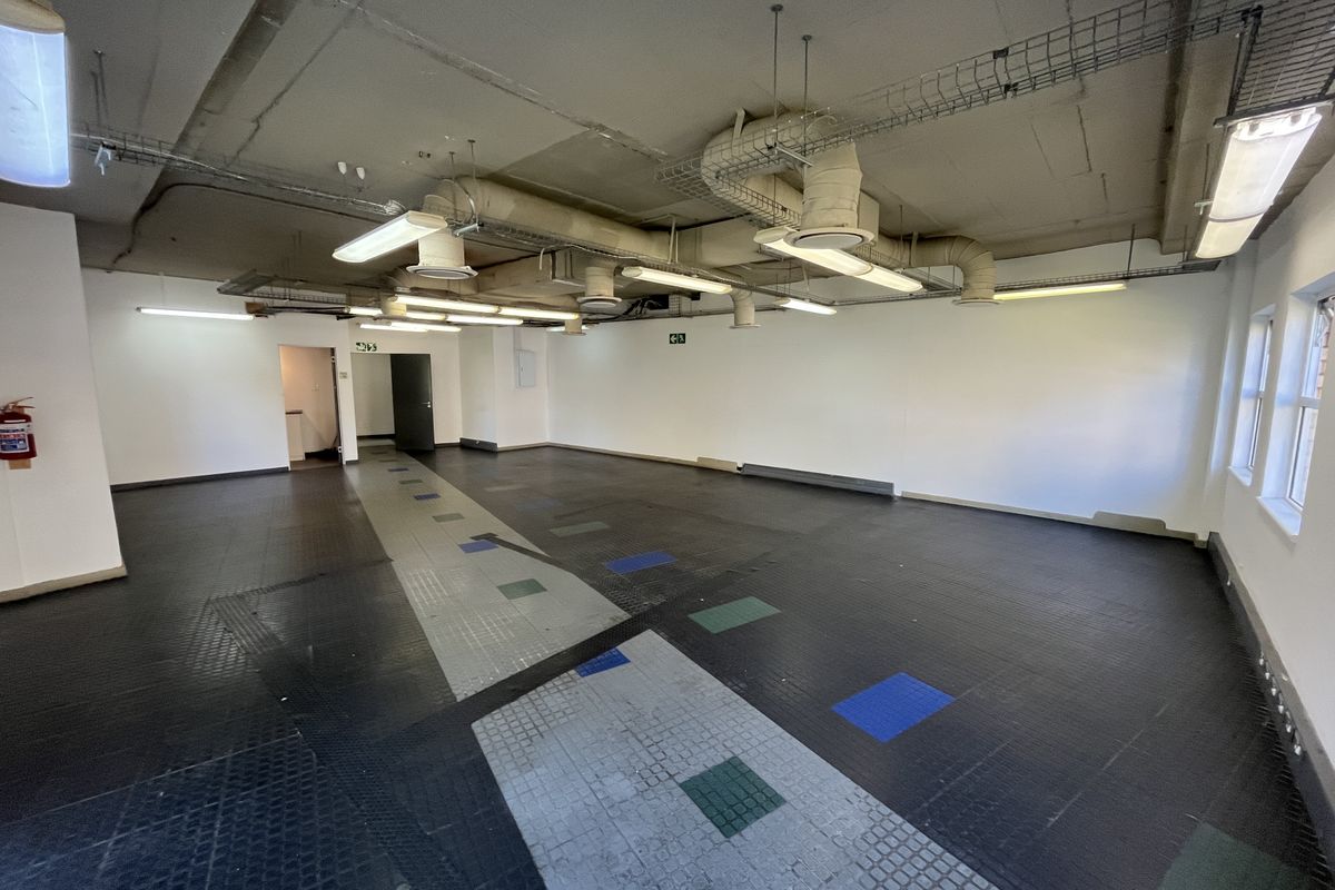 An open-plan office space featuring exposed industrial ceiling ducting, fluorescent lighting, and durable rubberized flooring, suitable for a variety of commercial configurations.
