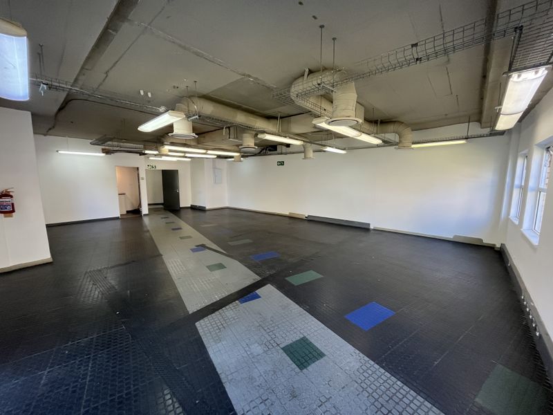 An open-plan office space featuring exposed industrial ceiling ducting, fluorescent lighting, and durable rubberized flooring, suitable for a variety of commercial configurations.