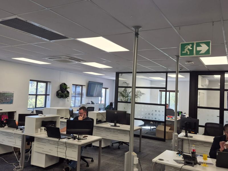 A modern open-plan office space featuring suspended ceilings, recessed lighting, air conditioning units, and glass partition walls with carpeted flooring.