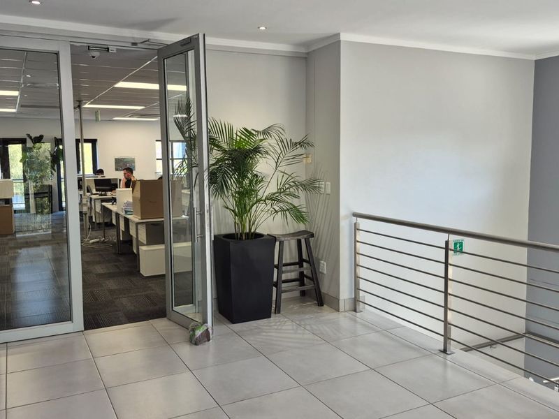 A bright office entrance featuring a glass door, tiled landing area, and a modern stainless steel balustrade overlooking the lower level.