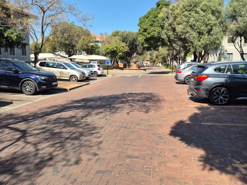 An outdoor parking area featuring brick paving, marked bays, and mature trees within an office park setting.