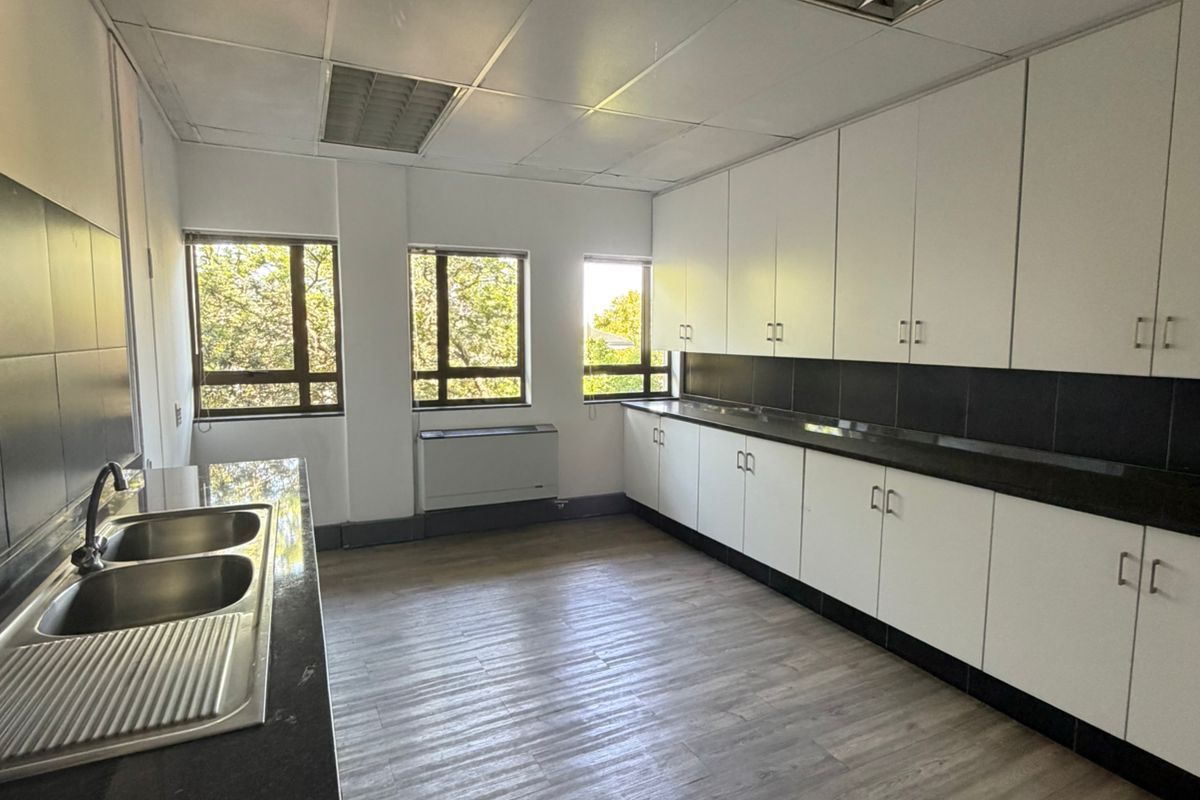 A functional office kitchenette featuring white cabinetry, dark countertops, double stainless steel sink, and wood-look vinyl flooring.