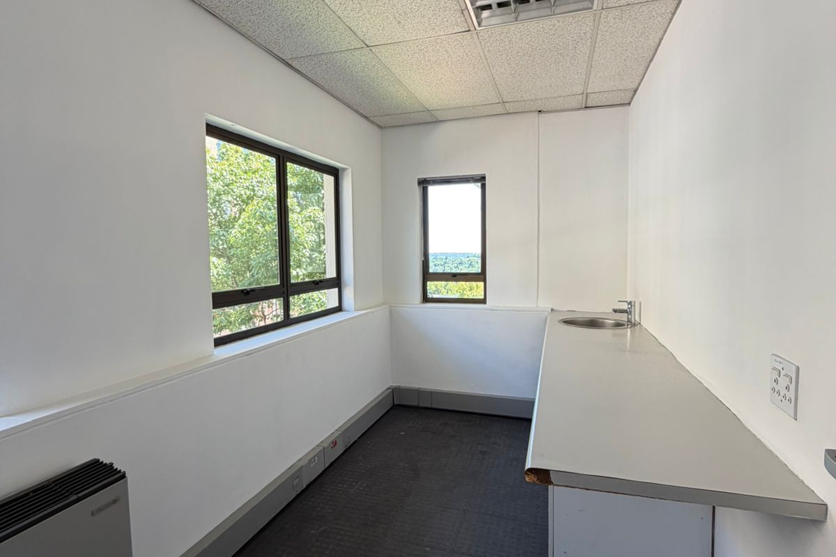 A small, empty office kitchenette featuring a stainless steel sink, laminate countertop, drop ceiling, and wall-mounted heater.