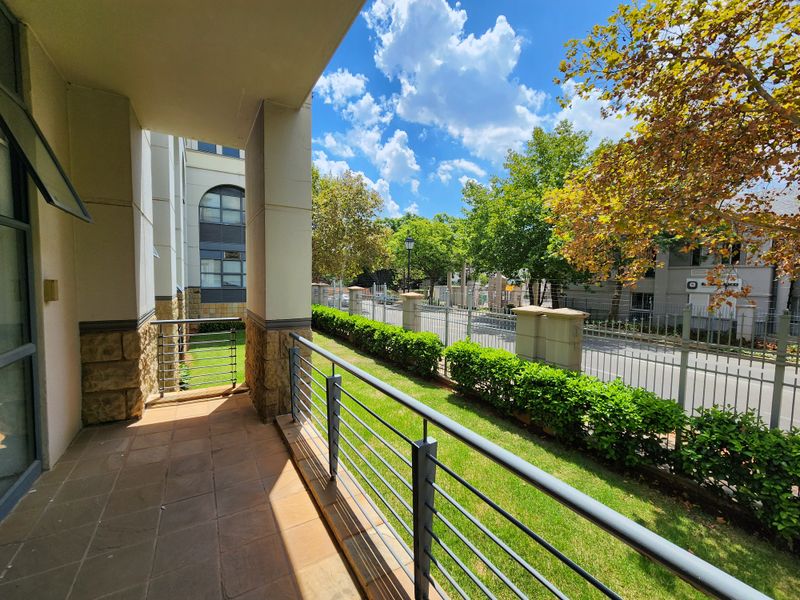 A covered ground-floor patio featuring tiled flooring, stone-clad pillars, and metal railings, overlooking a landscaped garden area and street-facing perimeter fence.