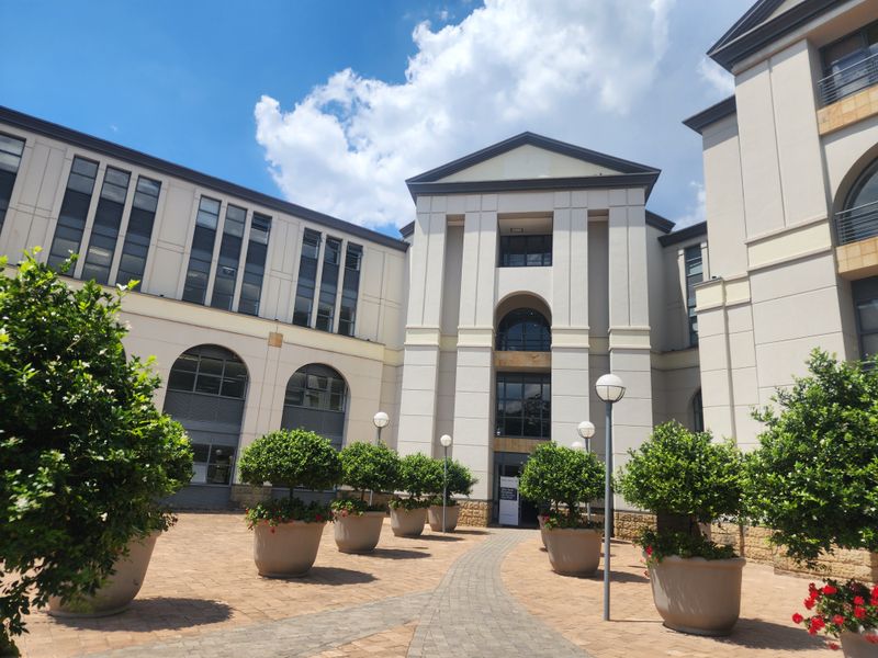 A well-maintained office building courtyard featuring paved walkways, architectural arched windows, and a multi-story facade with a central pediment entrance.