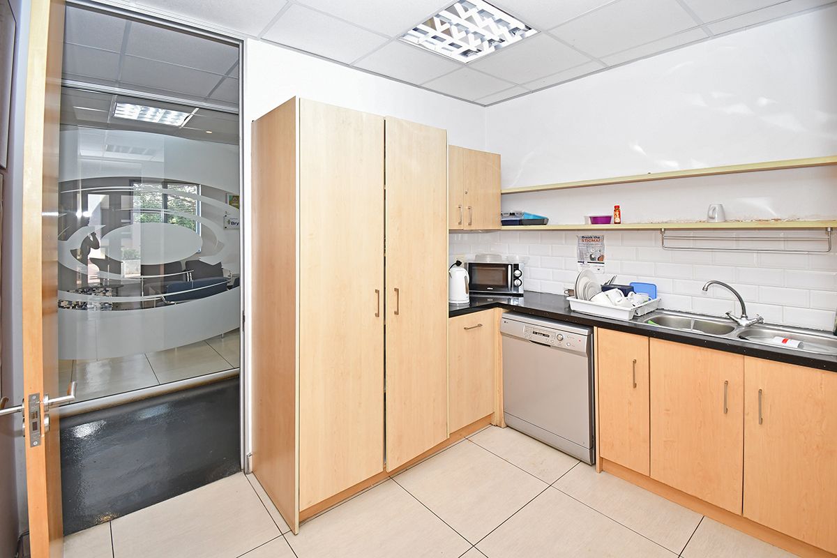 A functional office kitchenette featuring light wood cabinetry, a stainless steel sink, tiled backsplash, and integrated dishwasher, adjacent to a glass-partitioned office space.