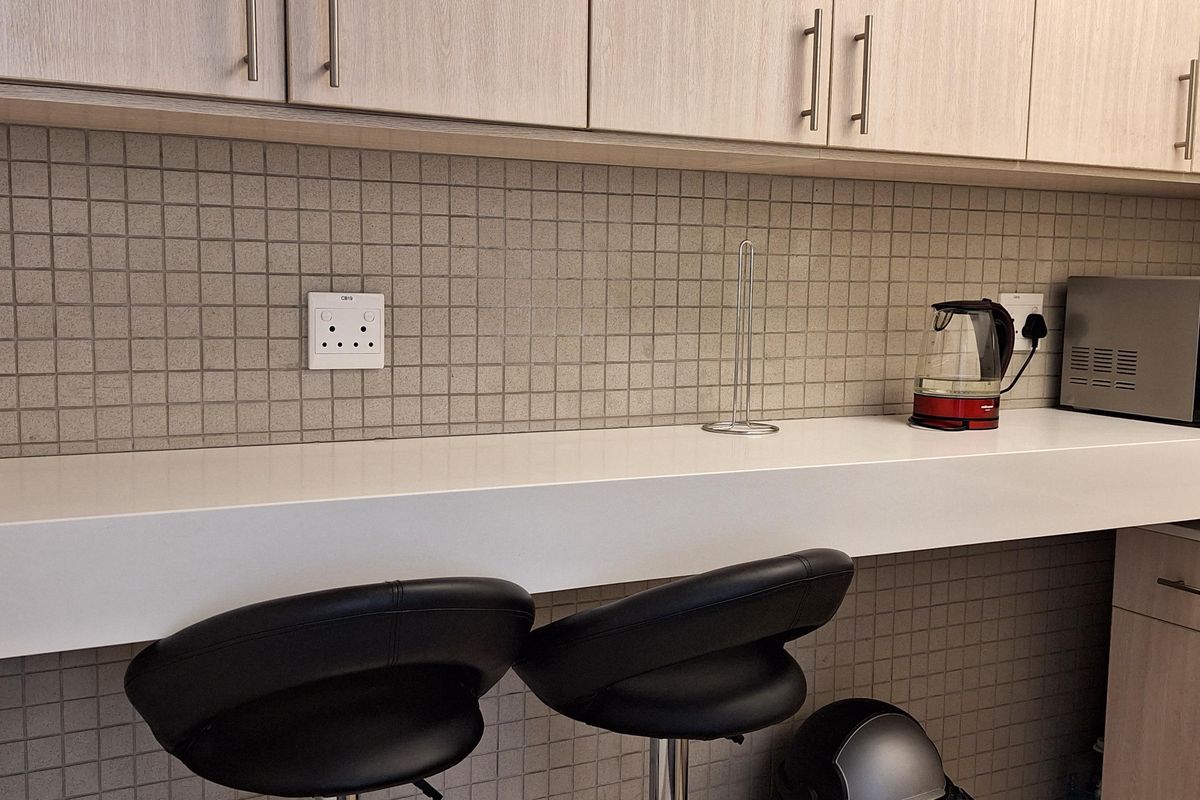 A functional office kitchenette area featuring light wood cabinetry, a white countertop, and a tiled backsplash with integrated electrical outlets.