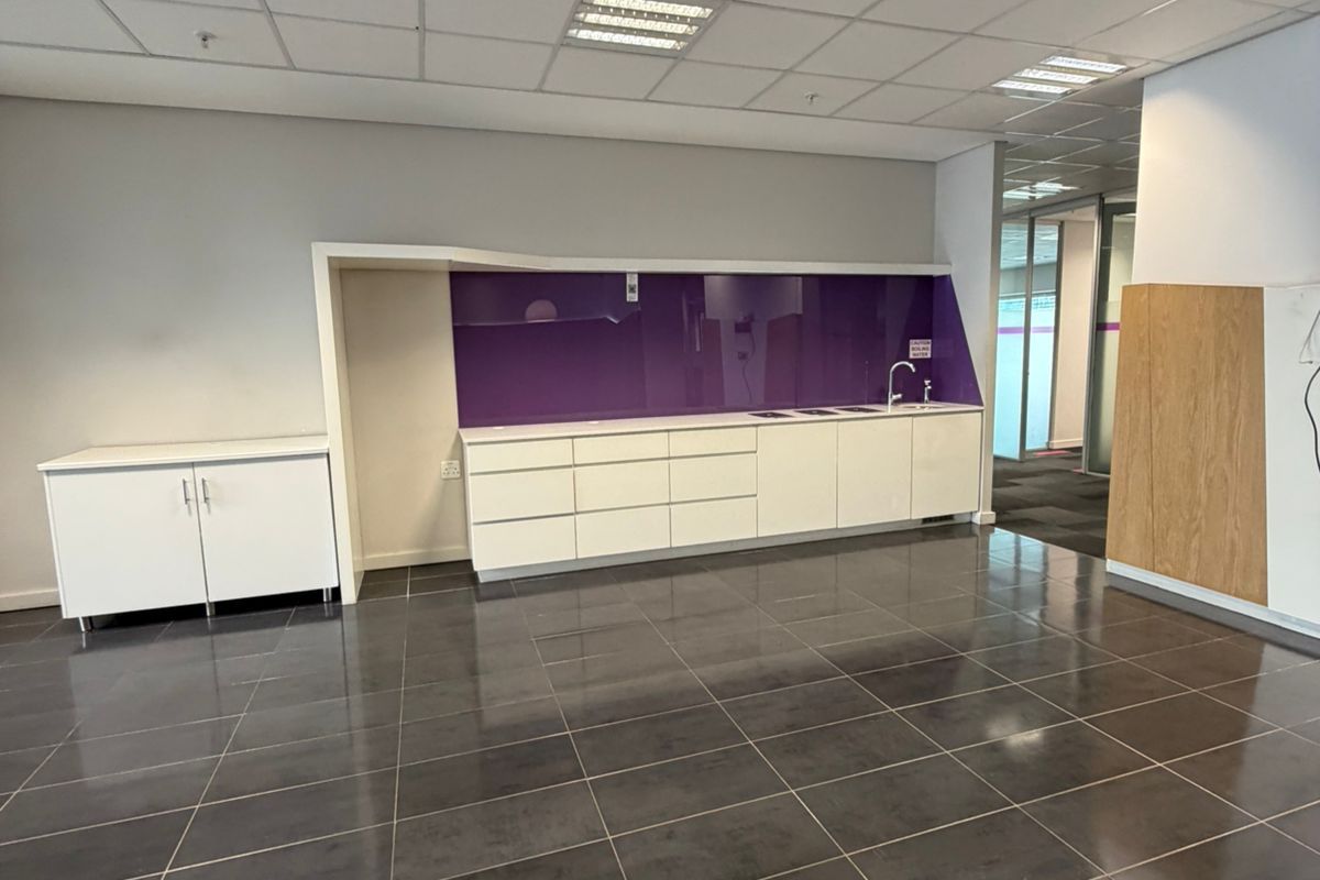 A modern office kitchenette area featuring white cabinetry, a purple glass splashback, integrated sink, and dark tiled flooring with a suspended ceiling.