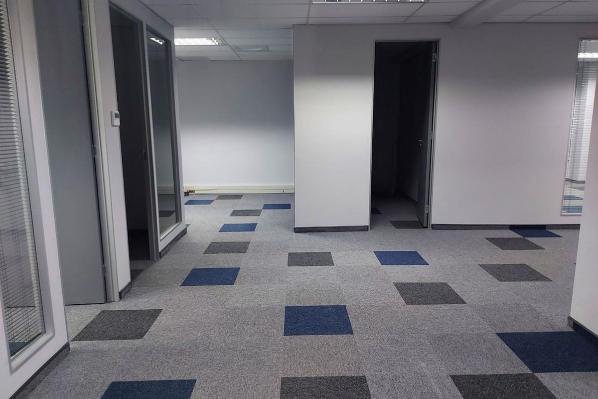 An empty office space featuring modern carpet tiles, glass partition doors, and a suspended ceiling with recessed lighting.