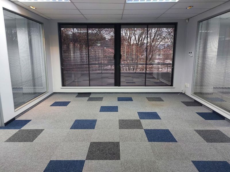 An empty office space featuring glass partitions, carpet tiles, and a suspended ceiling with recessed lighting, offering a professional work environment.