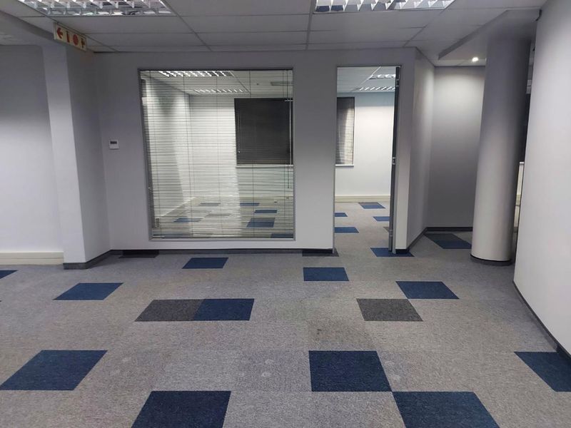An empty office space featuring carpet tiles, a suspended ceiling with recessed lighting, and a glass partition wall with integrated blinds.