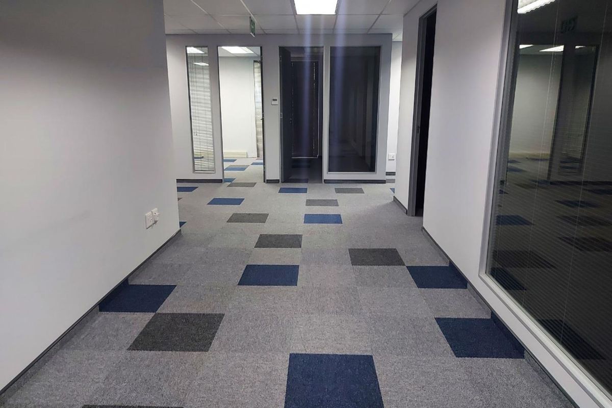 A clean, modern office hallway featuring patterned carpet tiles, suspended ceilings with recessed lighting, and glass partition walls leading to individual office spaces.