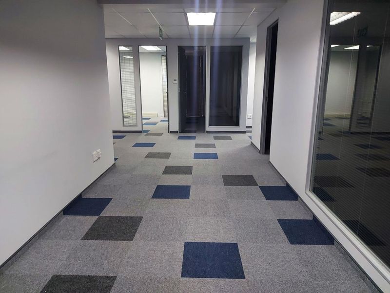 A clean, modern office hallway featuring patterned carpet tiles, suspended ceilings with recessed lighting, and glass partition walls leading to individual office spaces.