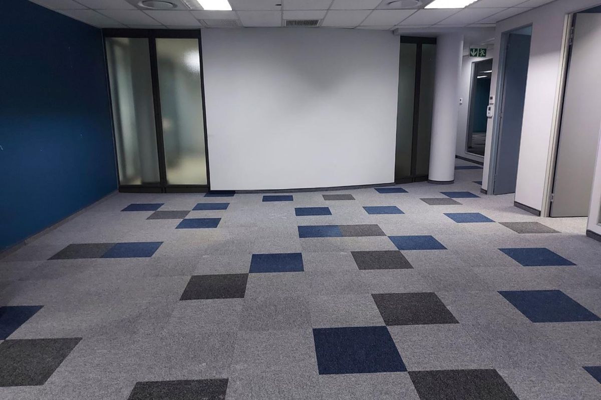 A spacious, empty office area featuring modern carpet tiles, suspended ceilings with recessed lighting, and frosted glass partitions for a professional workspace.