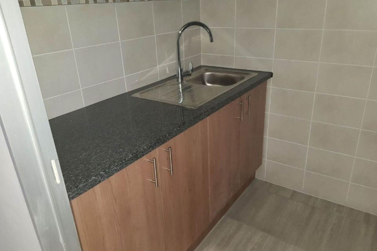 A compact kitchenette area featuring wooden cabinetry, a dark granite countertop, a stainless steel sink with a modern faucet, and tiled wall splashbacks.