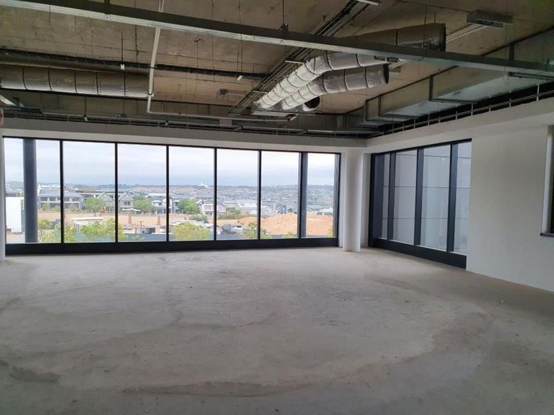 An expansive, open-plan office space in shell condition, featuring exposed industrial ceilings, concrete flooring, and large windows offering panoramic views.
