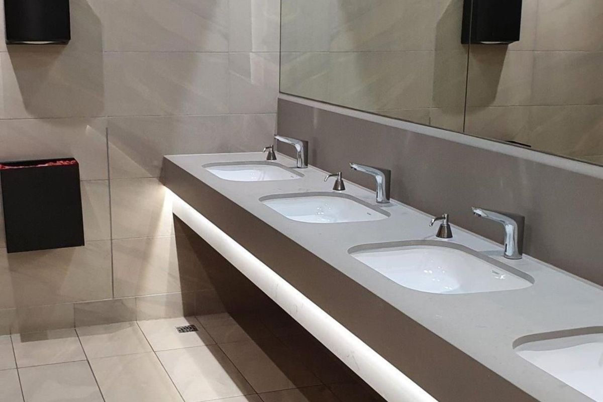 A modern, well-maintained commercial bathroom featuring a triple vanity basin with wall-mounted taps, large mirror, and integrated under-counter lighting.