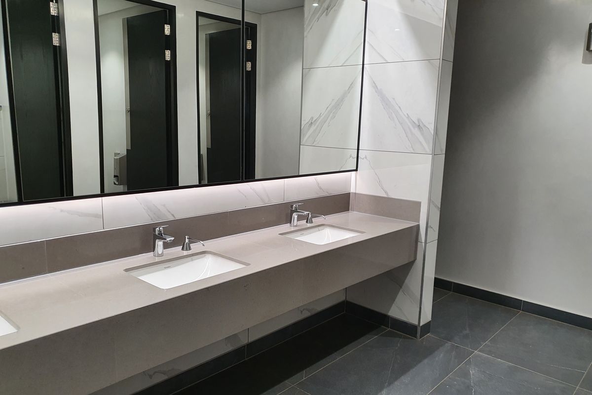 Modern office restroom featuring a dual-basin vanity with stone countertop, large wall-mounted mirror, and elegant marble-effect wall tiling.