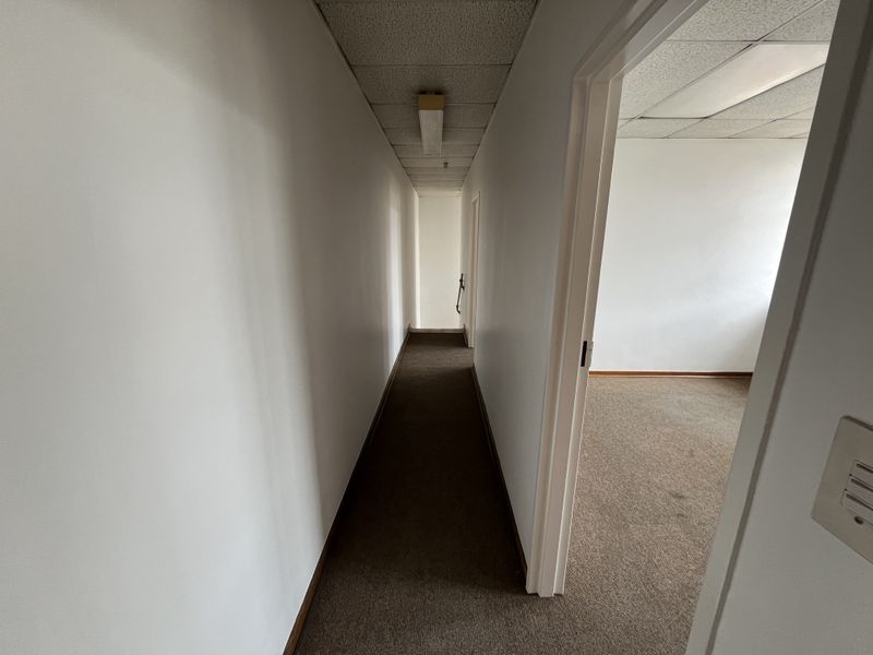 A narrow, empty office hallway featuring carpeted flooring and a suspended ceiling with integrated lighting, leading into an adjacent vacant room.
