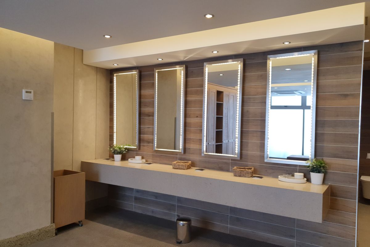 Modern office restroom vanity area featuring a long stone countertop, four integrated LED-lit mirrors, and wood-look wall tiling.