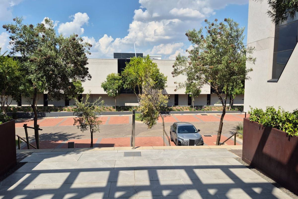 View from a paved terrace overlooking a modern office complex parking area with landscaped trees and structured building facades.