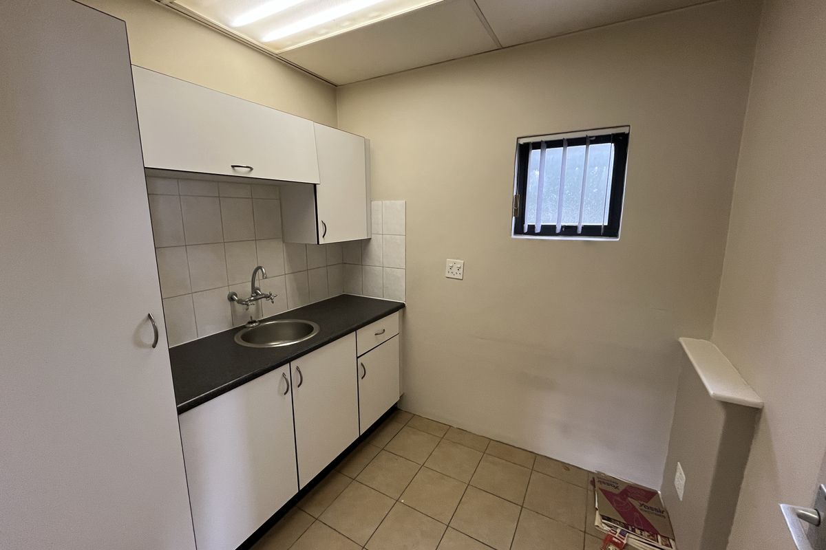 A compact office kitchenette featuring white built-in cabinetry, a stainless steel sink, tiled backsplash, and functional fluorescent lighting.