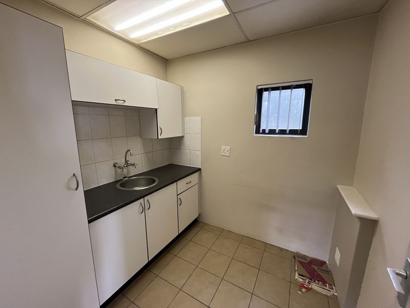 A compact office kitchenette featuring white built-in cabinetry, a stainless steel sink, tiled backsplash, and functional fluorescent lighting.