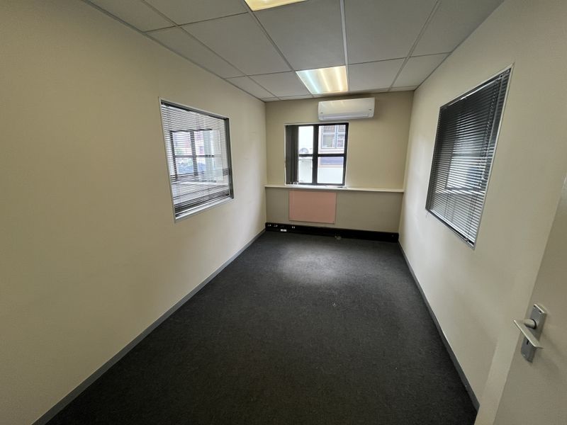 A vacant office space featuring carpeted flooring, a suspended ceiling with recessed lighting, and a wall-mounted split-system air conditioning unit.