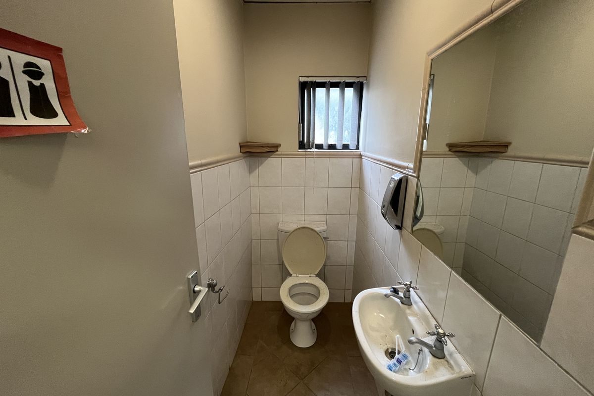 A functional office restroom featuring a wall-mounted basin, toilet, and tiled finishes, requiring cosmetic updates to improve its overall condition.