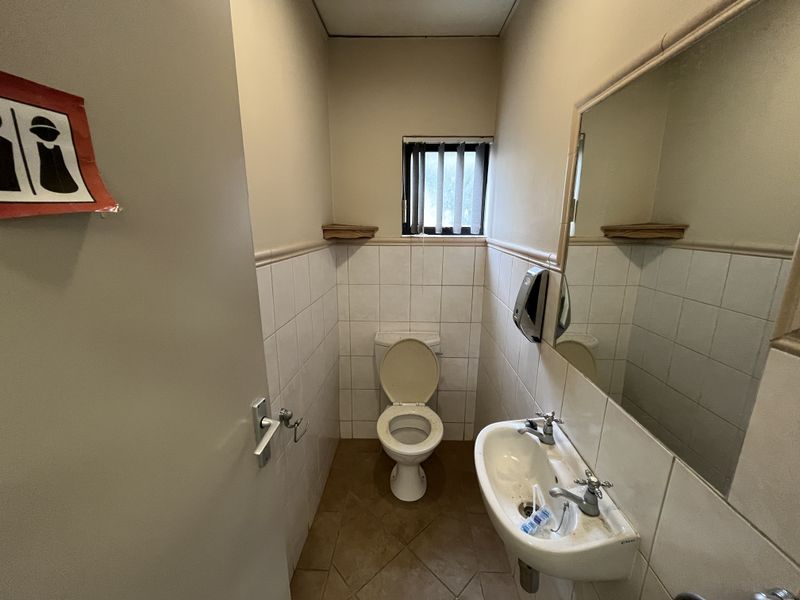 A functional office restroom featuring a wall-mounted basin, toilet, and tiled finishes, requiring cosmetic updates to improve its overall condition.