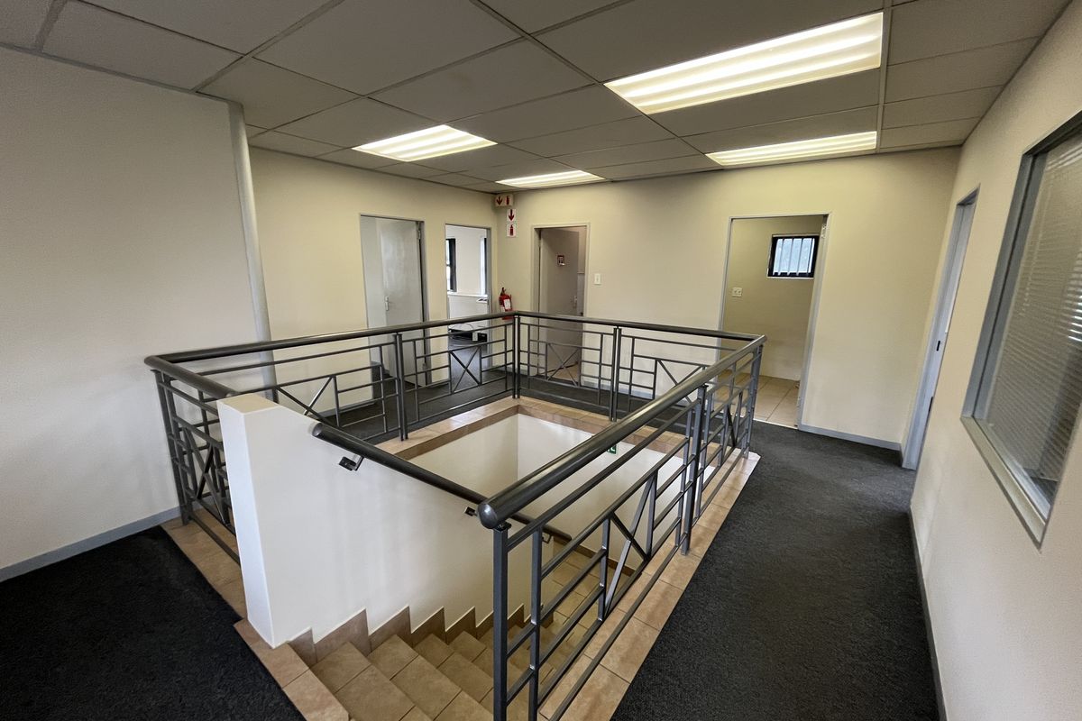 A spacious office building landing featuring a central staircase with metal balustrades, carpeted flooring, and recessed ceiling lighting leading to multiple office doorways.