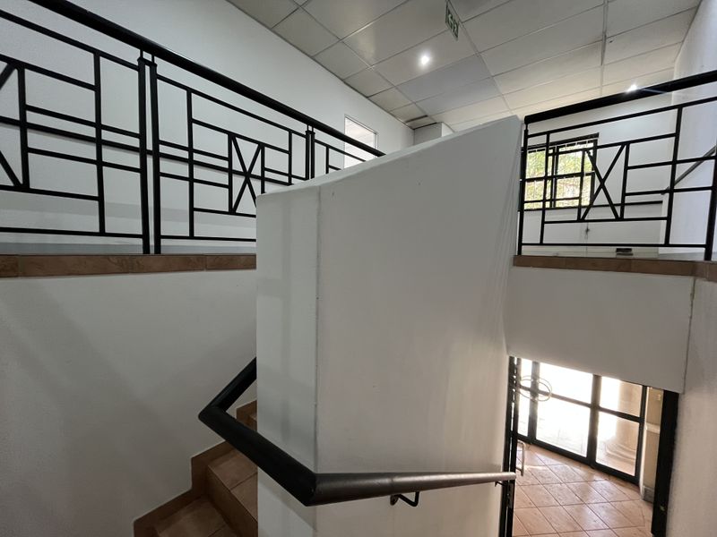 A functional office stairwell featuring tiled flooring, a metal balustrade, and a suspended ceiling with recessed lighting, providing access to the building entrance.