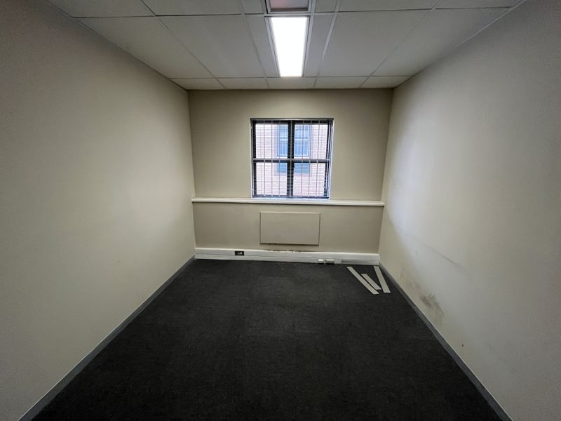 An empty office space featuring a suspended ceiling, recessed lighting, carpeted flooring, and a window with security bars.