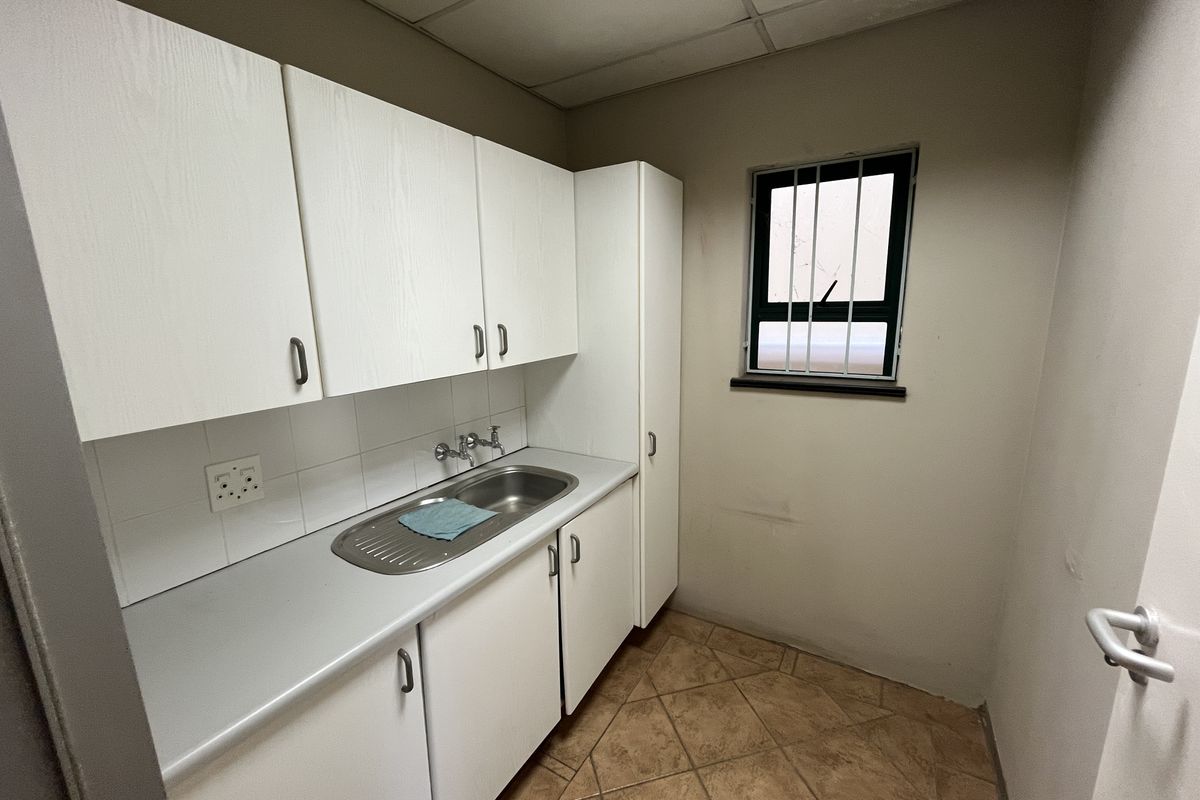 A compact kitchenette featuring white built-in cabinetry, a stainless steel sink, tiled splashback, and tiled flooring, suitable for office utility use.