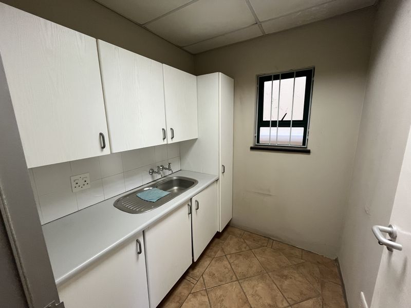 A compact kitchenette featuring white built-in cabinetry, a stainless steel sink, tiled splashback, and tiled flooring, suitable for office utility use.