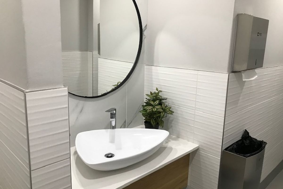 Modern office restroom featuring a contemporary vessel sink, wall-mounted faucet, circular mirror, and textured white wall tiling.
